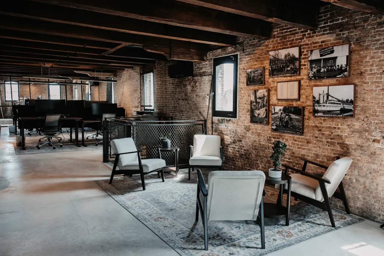 Het kantoor in Schiedam: witte fauteuils op een kleed voor een bakstenen muur met ingelijste zwart-witfoto's, met daarachter de werkplekken onder donkere houten balken.