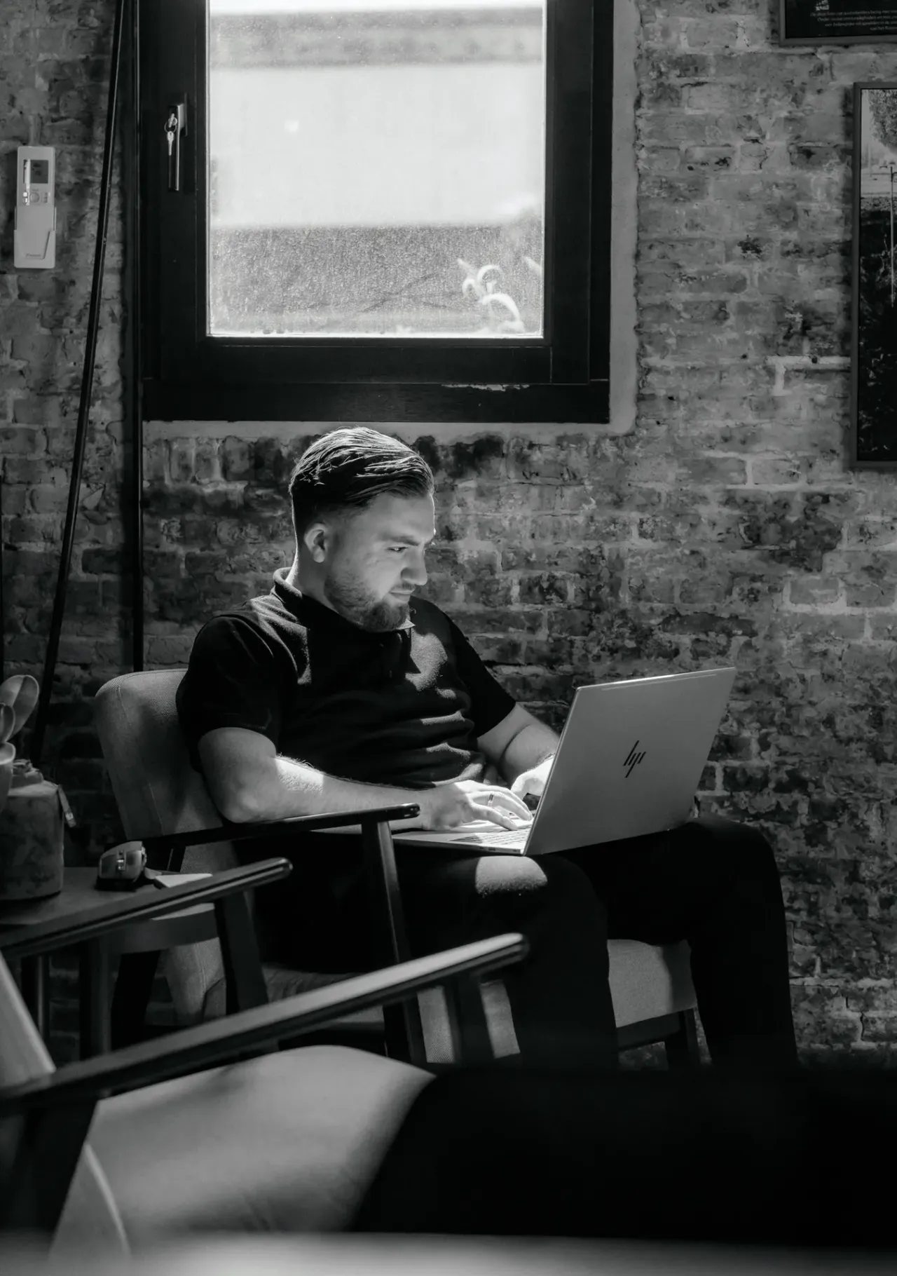Zwart-witfoto van een collega die in een fauteuil op een laptop werkt, voor een bakstenen muur onder een raam.