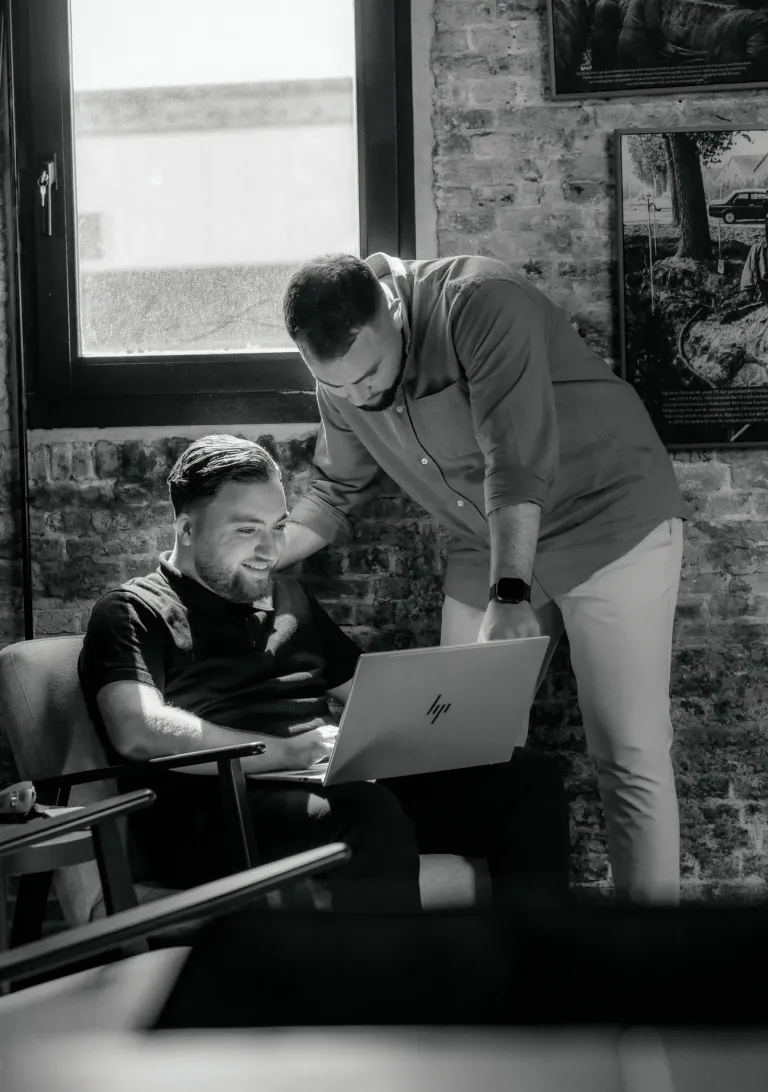 Zwart-witfoto op kantoor: een collega zit lachend met een laptop in een fauteuil, een tweede collega buigt zich over hem heen en kijkt mee. Op de achtergrond een bakstenen muur en een raam.